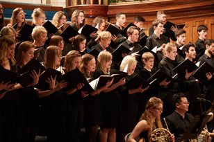 The choir performing on a stage, all dressed in black, accompanied by a brass section