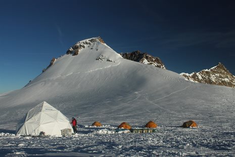 At the base of a snowy mountain are four orange tents and a white dome tent. A person in a red coat enters the dome tent.