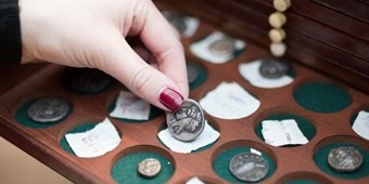 A hand reaches for an old coin, in a box of old coins. Each sits on its own green felt support