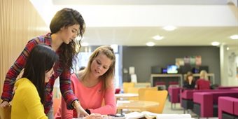 Three students look at papers on a table in the Humanities atrium