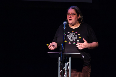 Sam Hirst stands at a podium and speaks into a microphone, wearing a shirt that says Don't Forget To Love Each Other, the text is surrounded by various pride flags.