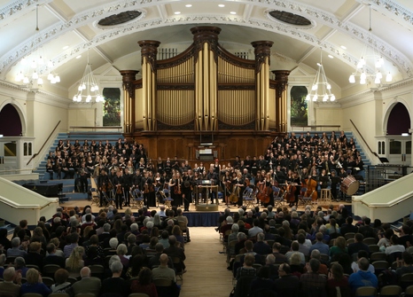 An orchestra play on a large stage with an organ behind them, to a large audience