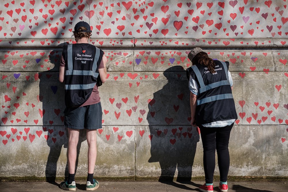 A wall covered in small painted red hearts, two people with their backs to the camera are adding hearts