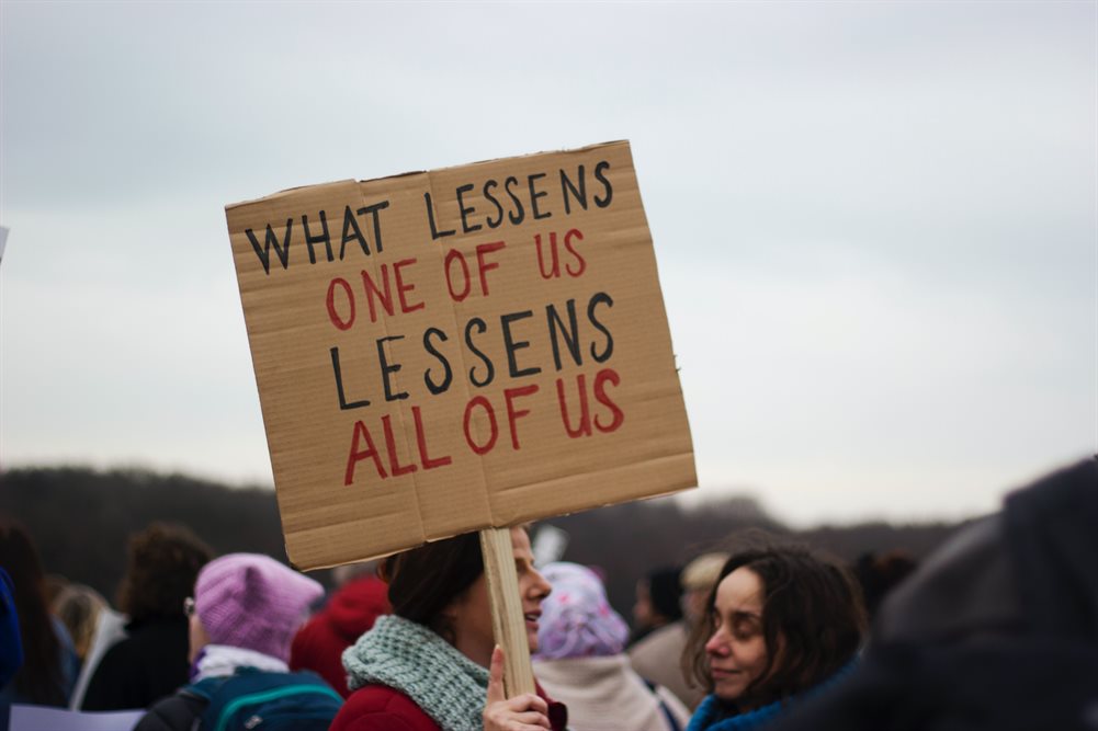 A person in a crowd holding up a sign which reads "What lessens one of us lessens all of us".