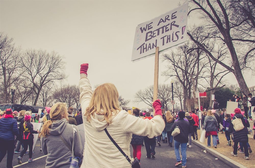 The back of a group of people during a demonstration. Focal point is a person raising their fist holding a sign which reads "we are better than this"