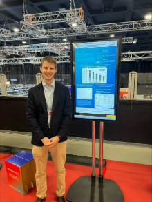 Photo of Chris Gilmartin at a conference, standing next to a poster displaying his research