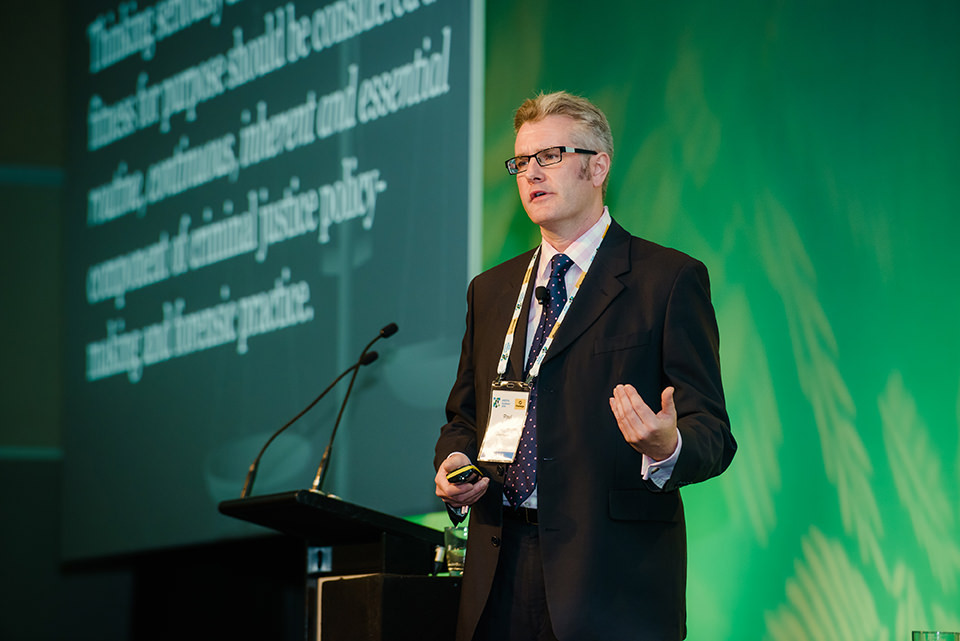 Professor Paul Roberts at a podium giving a presentation in a dark suit and navy blue spotted tie.
