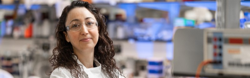 Head and shoulders portrait of researcher in white lab coat with equipment in background