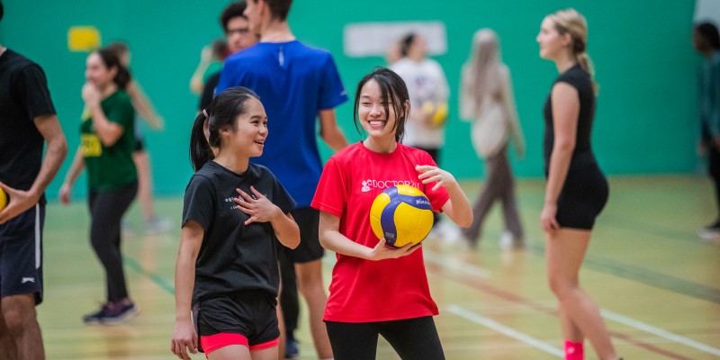 Two students chat in sports hall holding volleyball