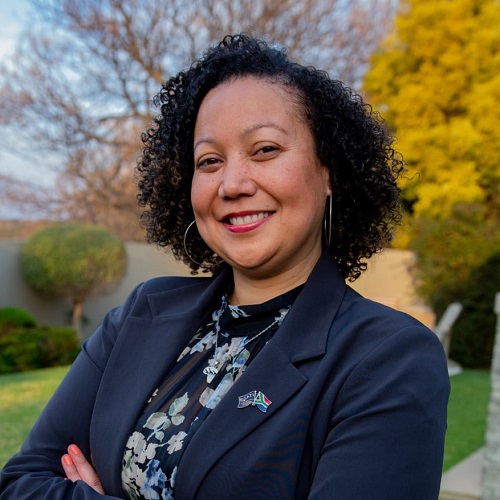 Photo of MBA student Mecia Peterson. Mecia is wearing a dark navy jacket and floral print top. Mecia is stood outside with trees in the background.