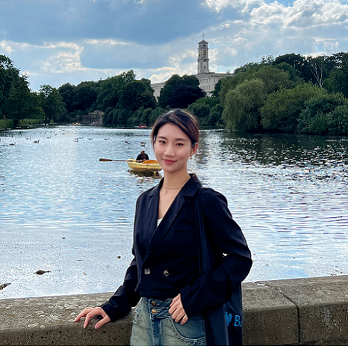 Chinese student Zixuan Xiao standing in front of the lake at University Park campus with the clock tower in the background. Wearing jeans and a dark blue jacket.