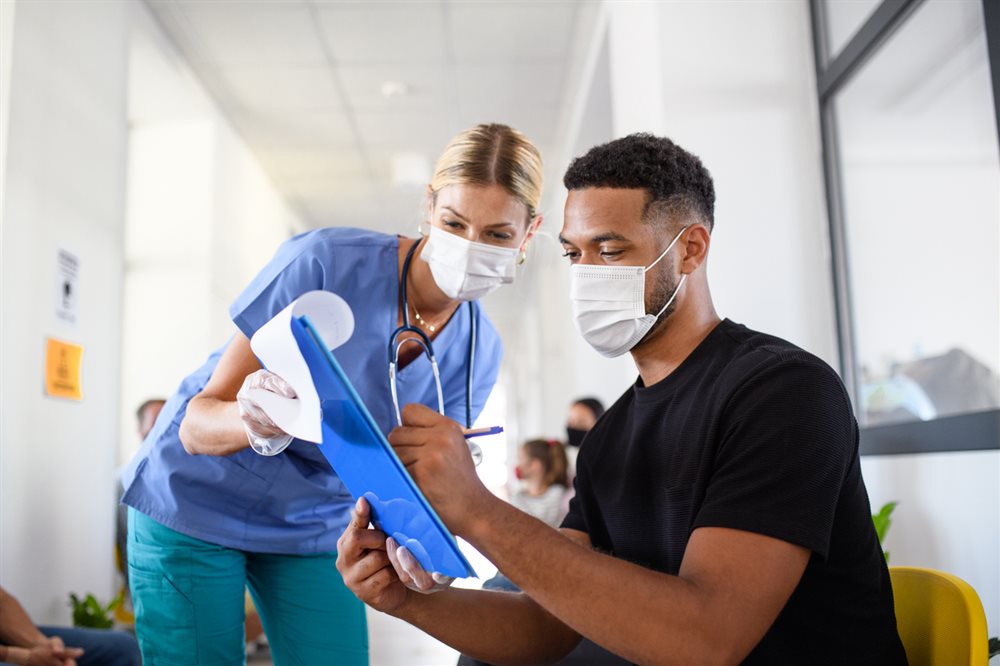 A medical person holding a clipboard out to a patient who is signing the paperwork on it.