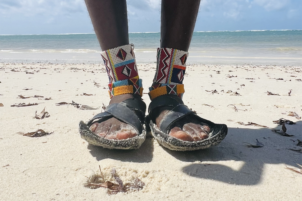 A pair of feet standing on a white sand beach wearing colourful cultural ankle bracelets.