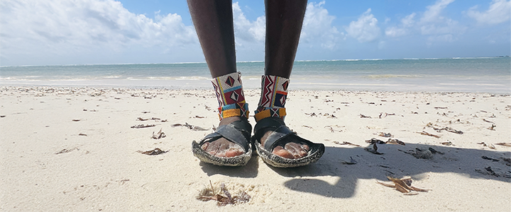 A pair of feet standing on a white sand beech wearing colourful cultural ankle bracelets.