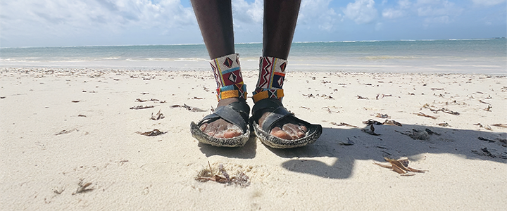 A pair of feet standing on a white sand beech wearing colourful cultural ankle bracelets.
