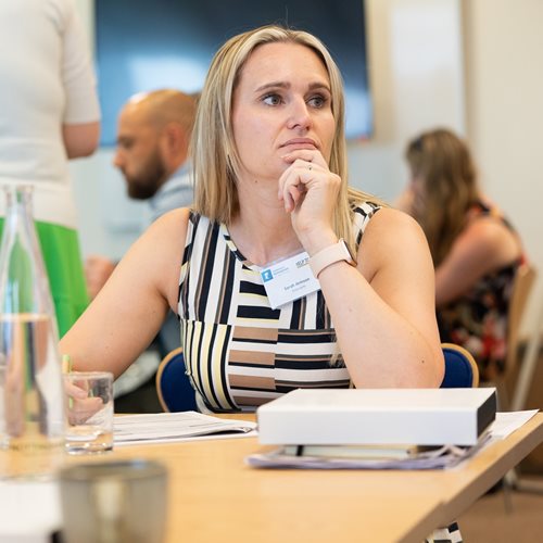 Photo of Sarah Armson, Commercial Manager of Doorcerts Manufacturing UK Limited. Sarah is sat in a conference room at a table. Sarah has blond shoulder length hair and is resting her chin on her hand, with her elbow resting on the table. In the background are other delegates attending the programme.