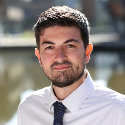 Photo of Jack Kavanagh. Jack has short dark hair and is wearing a white shirt with a blue tie. He is stood outside on Jubilee Campus with the lake in the background.