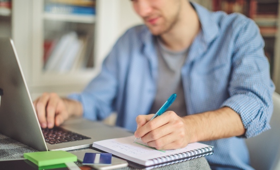 Male making notes while using a laptop