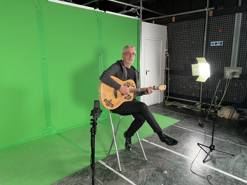 Person with guitar sitting on high stool in front of a green screen