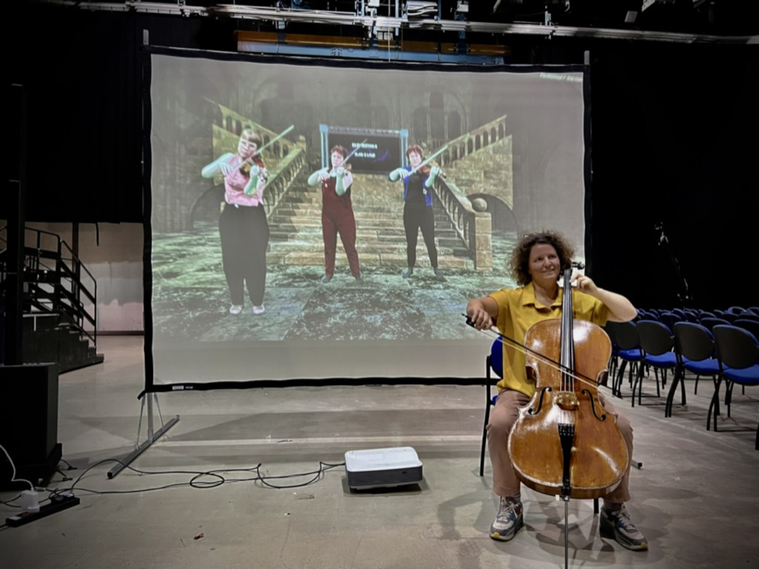 Person playing cello in front of a large screen showing three people playing violins