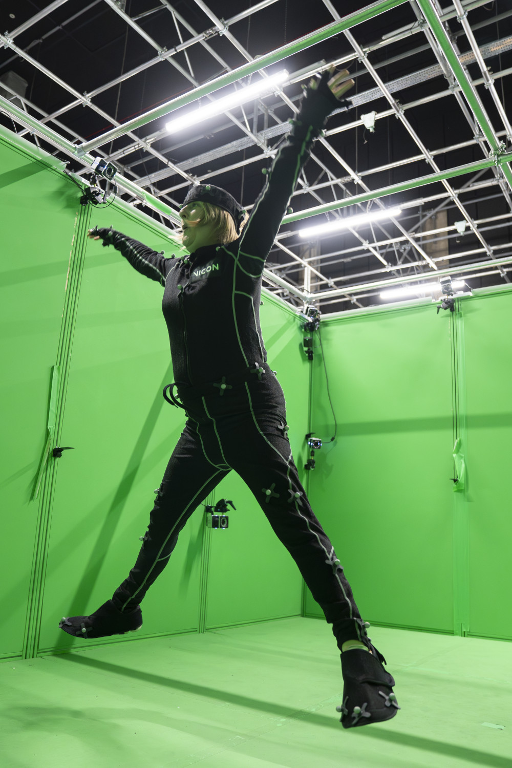 A woman in a back suit jumping up with her arms out. She is in front of a green screen in a film studio