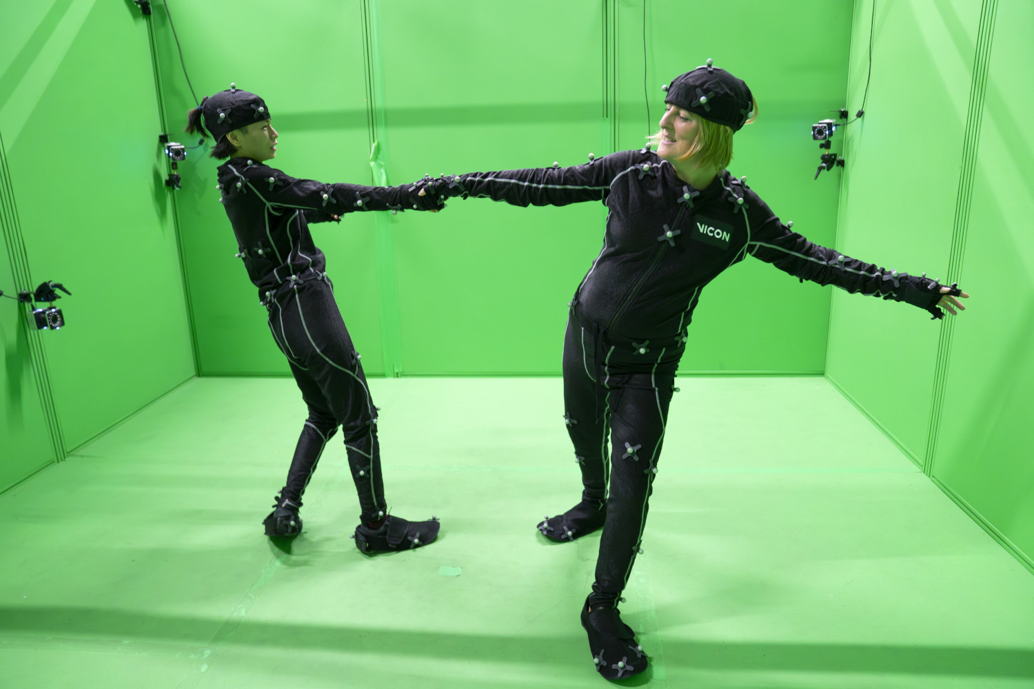 Two women wearing back suits holding hands and leaning away from each other. They are in front of a green screen.