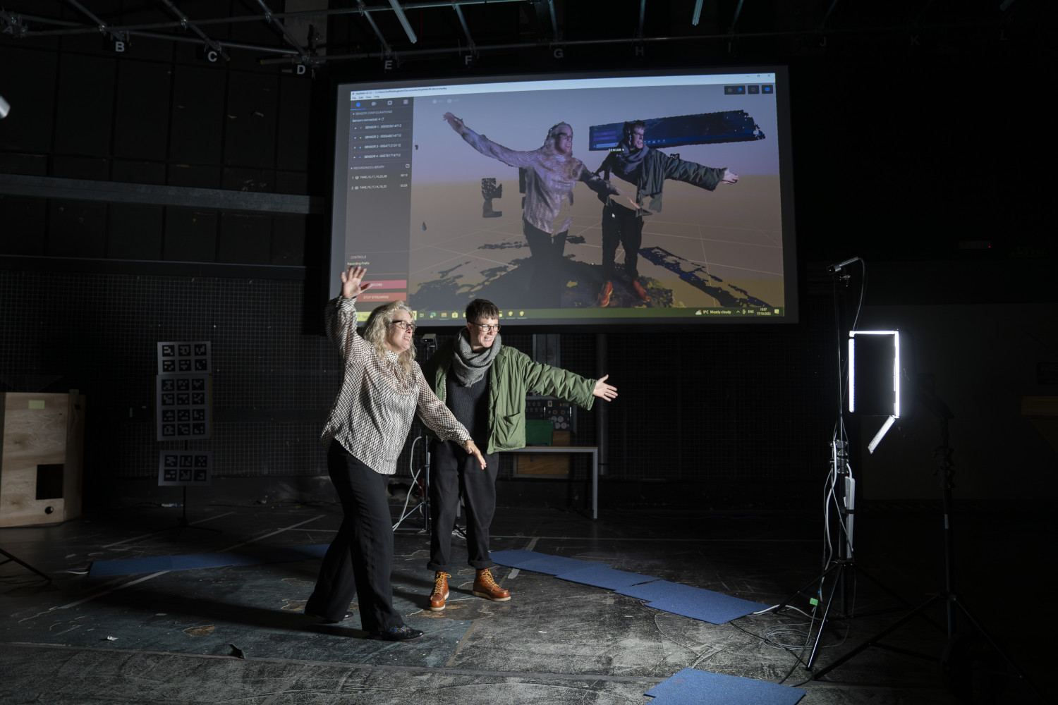 Two women stand in front of a projector screen holding their arms open – their image is shown on the projector screen.