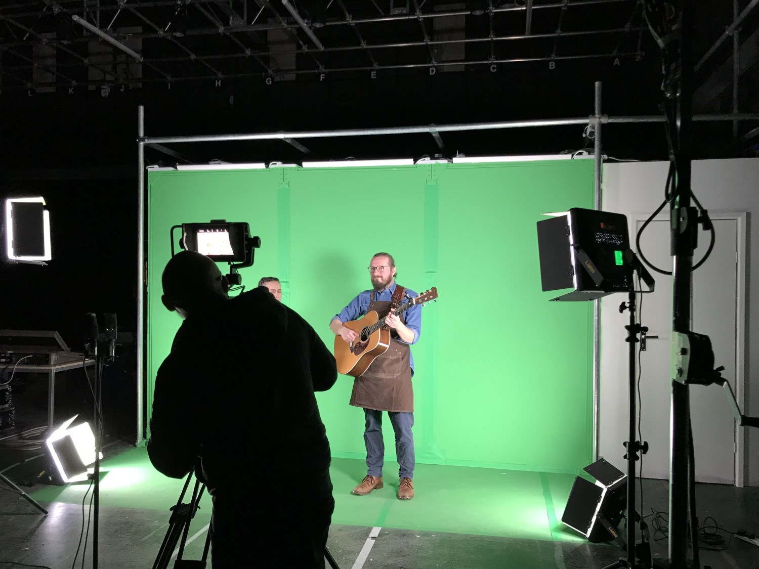 Person playing guitar standing in front of green screen while another person stands in front filming them with a camera on a tripod