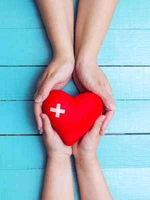 Two pairs of hands holding a red heart with a white cross on it