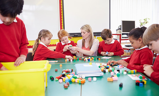 Female primary school teacher sitting at table with male and female pupils counting blocks