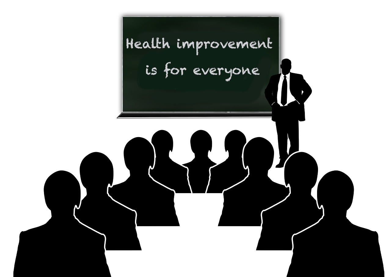 An image of doctor standing in front of group of people and by a board that reads 'Health improvement is for everyone'.