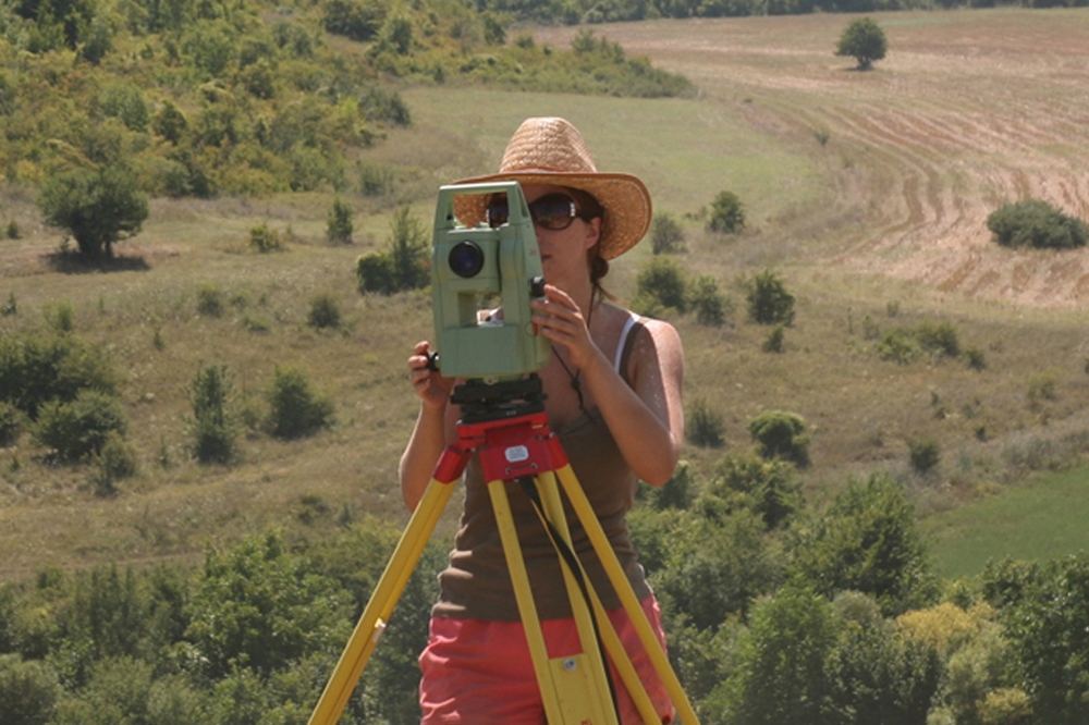 A female student taking part in a fieldwork project, measuring the land, wearing sunglasses and a sun hat.