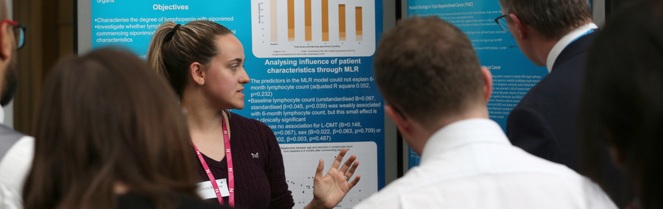 Clinical Academic Fellow standing in front of their research poster talking about their research to interested listeners.