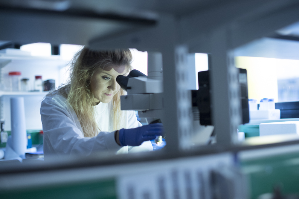 Female postgraduate student in a lab coat looking through a microscope studying cancer cells