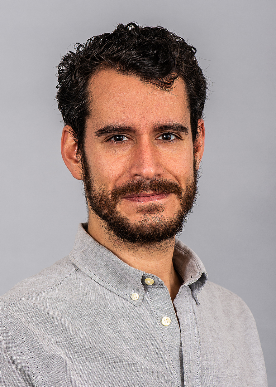 A professional headshot of a young man with dark hair and beard, wearing a grey shirt