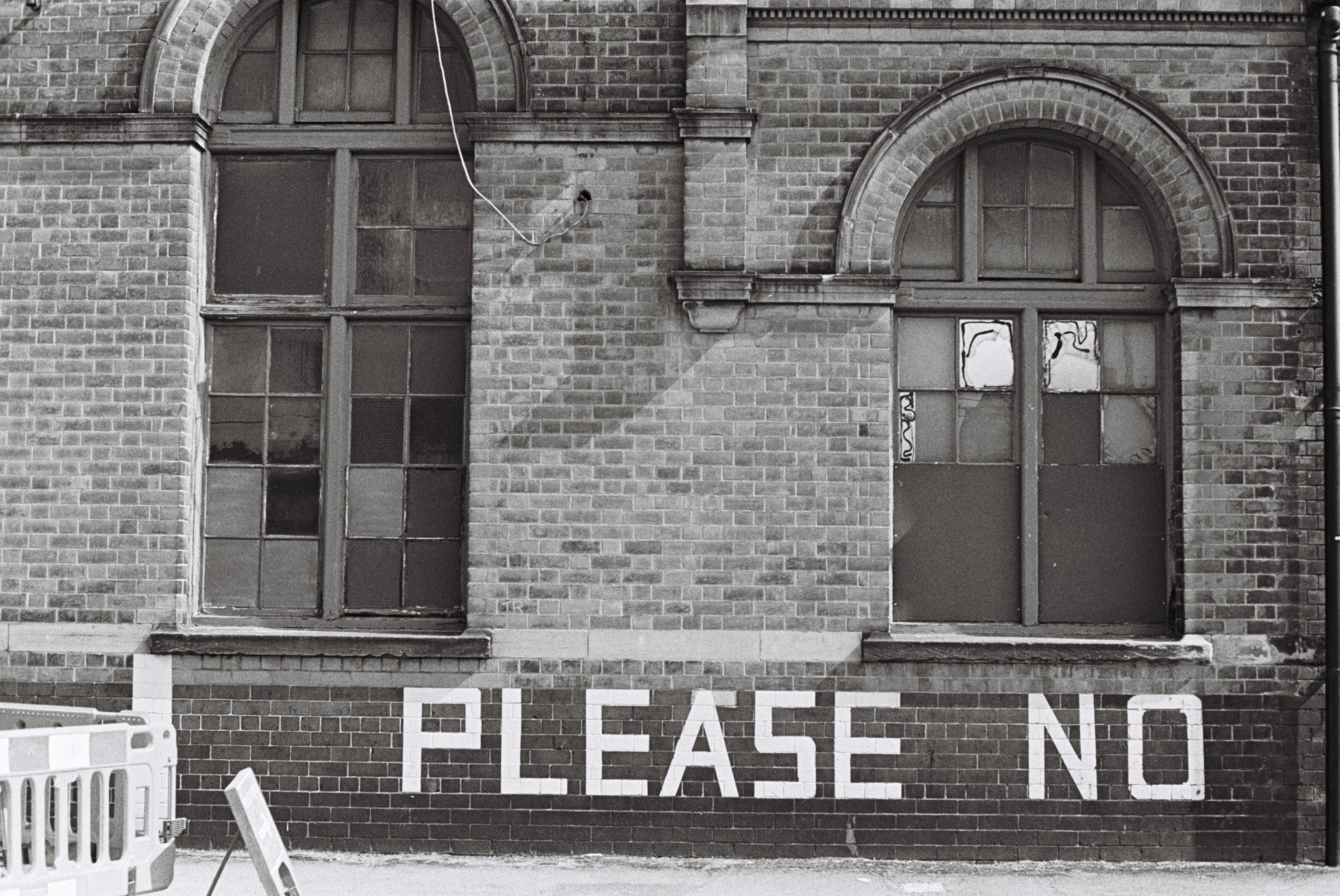 Black and white photograph of two large windows in a brick building with 'PLEASE NO' written in white on the wall below.