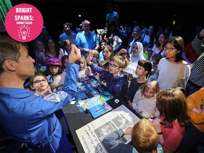 White man in blue boiler suit presenting to a large diverse crowd of children and adults