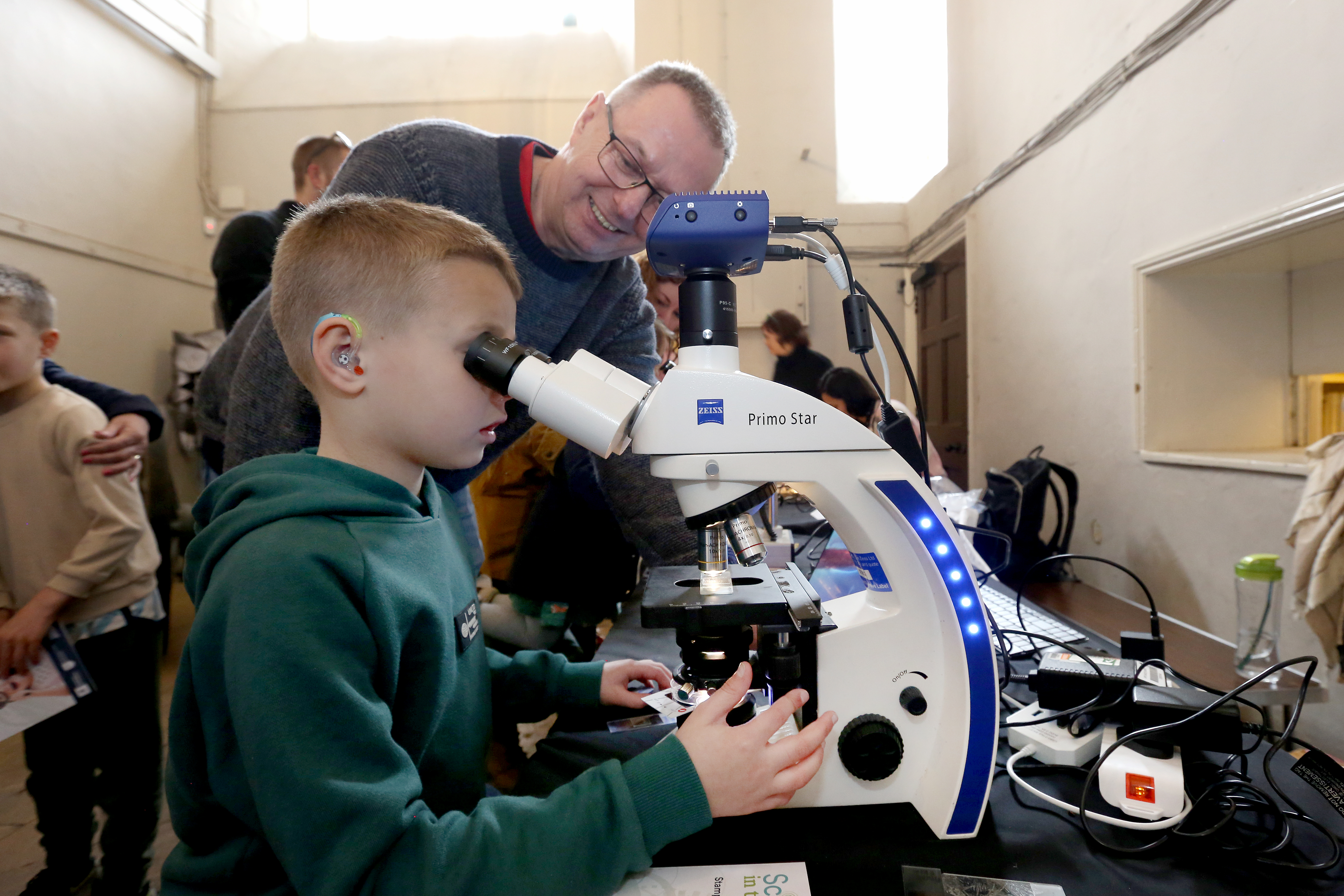 A small boy looking into a huge telescope. There is an older man beside him smiling