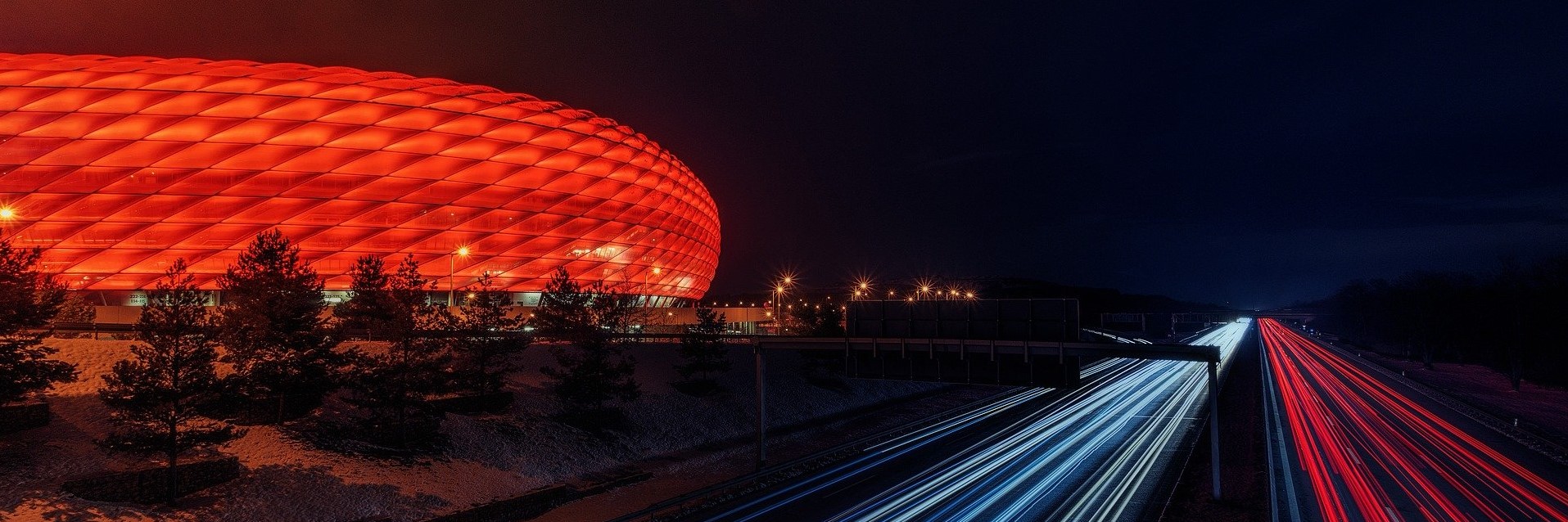 A football stadium lit up in red at night next to a fast moving road