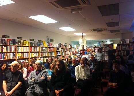 Room full of people listening to a speaker at the front, with bookcases lining the walls