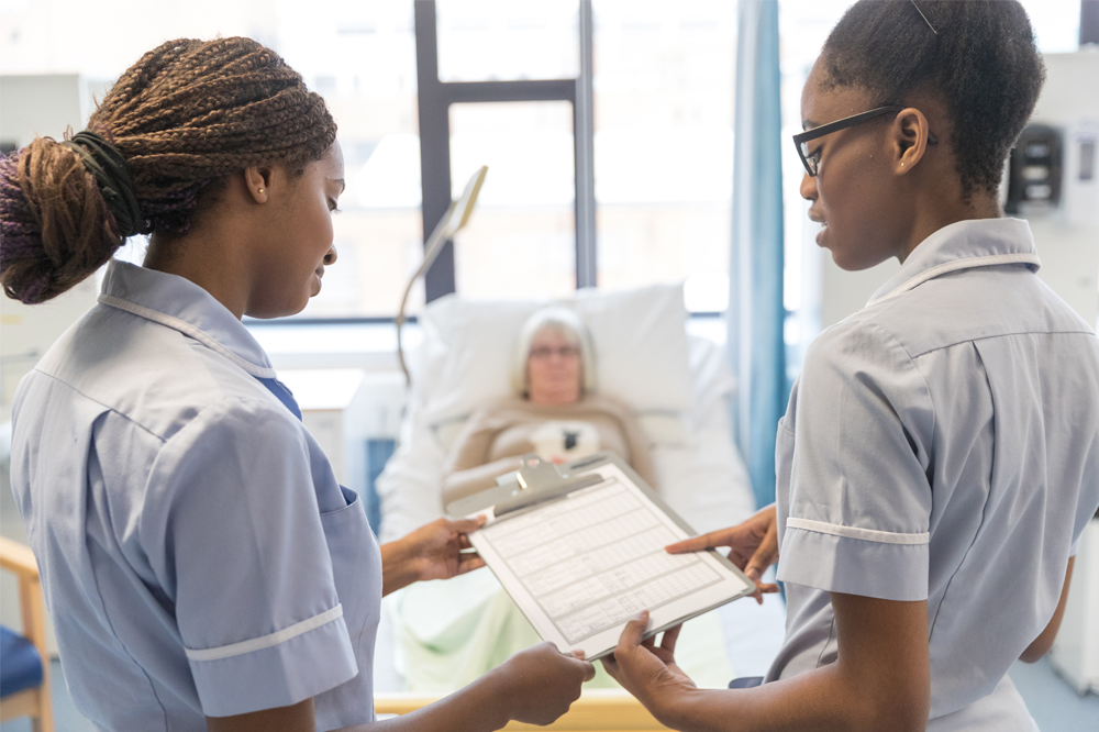 Two nursing students discussing a patient's notes in the Clinical Skills Centre. The patient is in a hospital bed in the background.